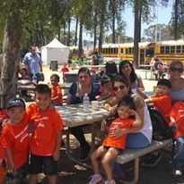 A group of adults and children in orange shirts sit at a picnic table under trees on a sunny day. A yellow school bus is visible in the background.
