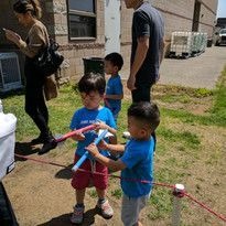 Children in blue shirts play outdoors near a building. They are holding paper, creating a playful and energetic atmosphere. Adults are present nearby.