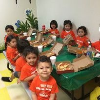 Children in orange shirts sit around a table with pizza and drinks in a bright room. The mood is cheerful and festive, with decorations on the wall.