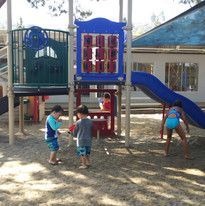 Children playing on a playground with a blue slide, climbing structures, and a shaded area. The scene conveys a joyful, sunny day.