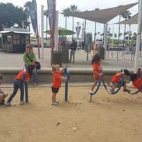 Children in orange shirts play on wavy metal bars at a park. A few adults supervise while others stand nearby. The scene is lively and joyful.