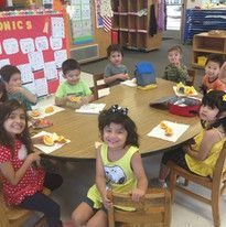 A group of young children sit smiling around a round table in a classroom, enjoying sliced apples. The atmosphere is cheerful and welcoming.