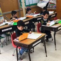 Children sitting at desks in a classroom, focused on writing in notebooks. The setting is studious, with bright folders and classroom decor visible.