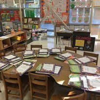 A lively classroom with a round table filled with colorful worksheets, surrounded by chairs. Bright posters and books line the walls, creating an engaging atmosphere.