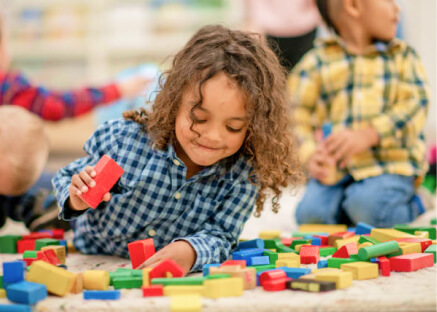 A child in a blue checkered shirt plays with colorful building blocks on the floor, smiling. Other children in the background are also engaged in play.