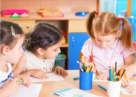 Three young children are sitting at a table in a classroom, focused on drawing with colored pencils. The atmosphere is cheerful and collaborative.