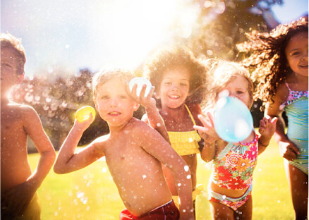 Children joyfully play on a sunny day, splashing water and holding balloons. The scene is vibrant, filled with sunlight and laughter.