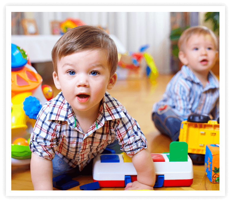 Two toddlers in plaid shirts play on a wooden floor with colorful toys, including blocks and a truck, conveying curiosity and joy.