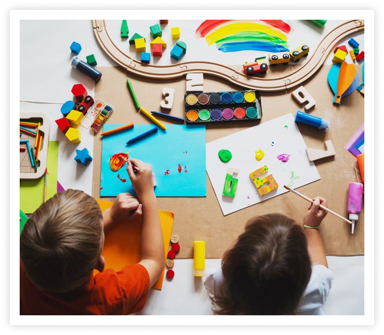 Two children are engaged in painting at a table filled with colorful art supplies, wooden blocks, and a toy train. A bright, creative atmosphere prevails.