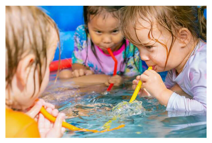 Three young children playfully use colorful straws to blow bubbles in a small pool, creating splashes. Their expressions are focused and joyful.